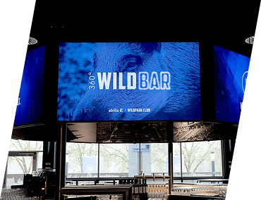 Ein großer digitaler Bildschirm im Inneren einer modernen Bar zeigt 360° WILDBAR mit einem Bild eines Wildschweins im Hintergrund. Der Barbereich darunter ist mit Glaswaren und Fenstern ausgestattet, die Bäume im Freien zeigen.