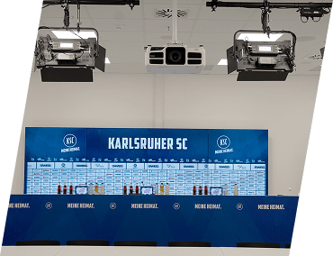 Ein Pressekonferenzraum mit einem großen blauen KARLSRUHER SC-Hintergrund, mehreren Mikrofonen, Wasserflaschen auf einem Tisch, Deckenbeleuchtung und sichtbaren Kameras.
