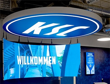 Ein blaues, rundes Schild mit dem weißen KSC-Logo hängt von der Decke herab. Darunter zeigt ein digitaler Bildschirm in fetten weißen Buchstaben WILLKOMMEN an. Ein weiterer Bildschirm mit Informationen ist an der Wand im Hintergrund zu sehen.