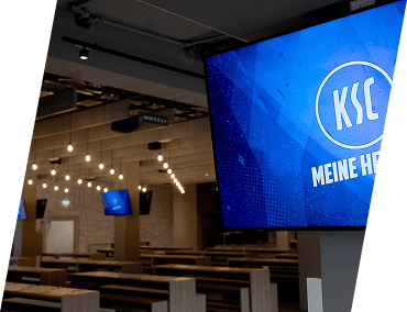 Eine moderne, leere Cafeteria mit großen Holztischen und Hängelampen. Auf mehreren Flachbildfernsehern ist ein blauer Bildschirm mit KSC MEINE HE teilweise sichtbar. Die Atmosphäre wirkt sauber und modern.