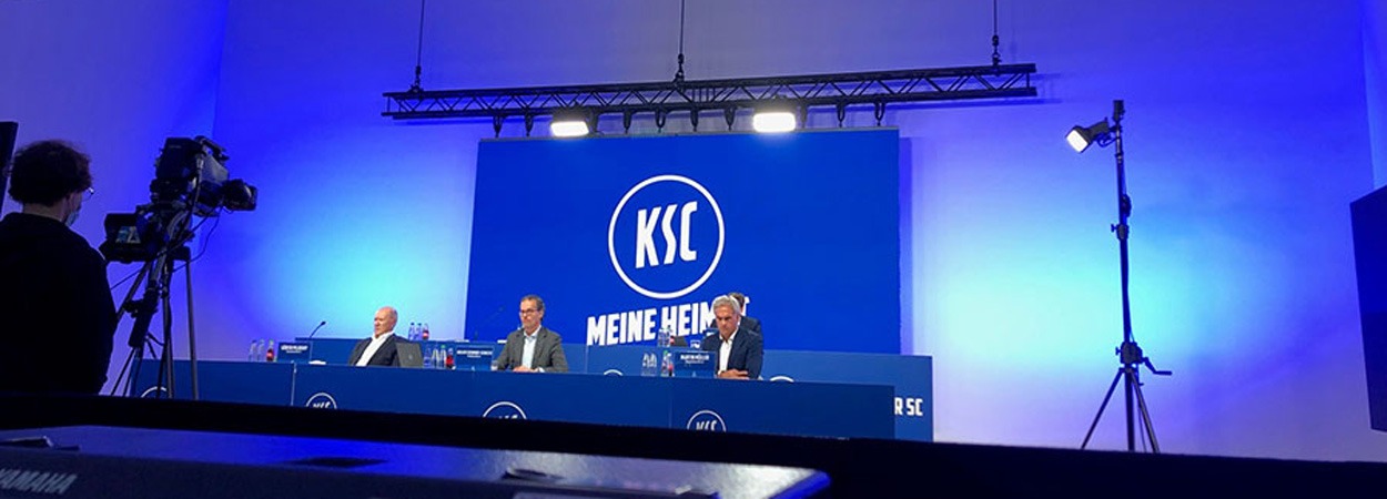 Drei Männer sitzen an einem langen blauen Tisch auf einer Bühne, hinter ihnen ein großer Bildschirm mit dem KSC-Logo und "MEINE HEIMAT". Der Raum ist hell erleuchtet, Kameras und Beleuchtungsanlagen sind zu sehen.