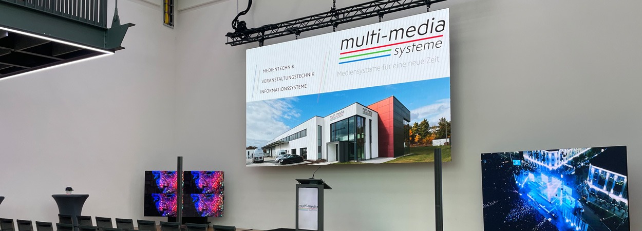 Ein großer Präsentationsbildschirm in einem modernen Veranstaltungsraum zeigt das Logo und das Bild von Multimedia Systeme mit einem Foto eines modernen Gebäudes. Darunter befinden sich ein Podium, Stehtische und kleinere digitale Bildschirme.