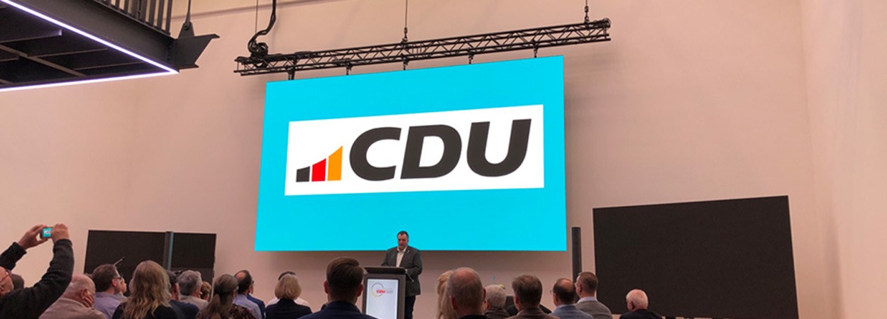 Ein Redner steht an einem Podium vor einem Publikum, dahinter ist auf einem großen Bildschirm das CDU-Logo in schwarzen Buchstaben auf blauem Hintergrund zu sehen.