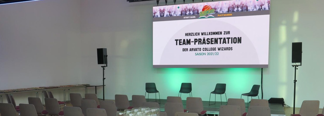 Ein Präsentations-Setup mit Stühlen vor einer Bühne, vier Stühlen und Mikrofonen an der Vorderseite und einer großen Leinwand mit der Aufschrift "Herzlich Willkommen zur Team-Präsentation der Arvato College Wizards Saison 2021/22.".