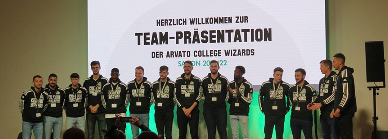 Ein Basketballteam steht in passenden Trainingsanzügen auf der Bühne vor einer großen Leinwand mit der Aufschrift "Team-Präsentation der Arvato College Wizards Saison 2021/22" und begrüßt die Teilnehmer der Team-Präsentationsveranstaltung.
