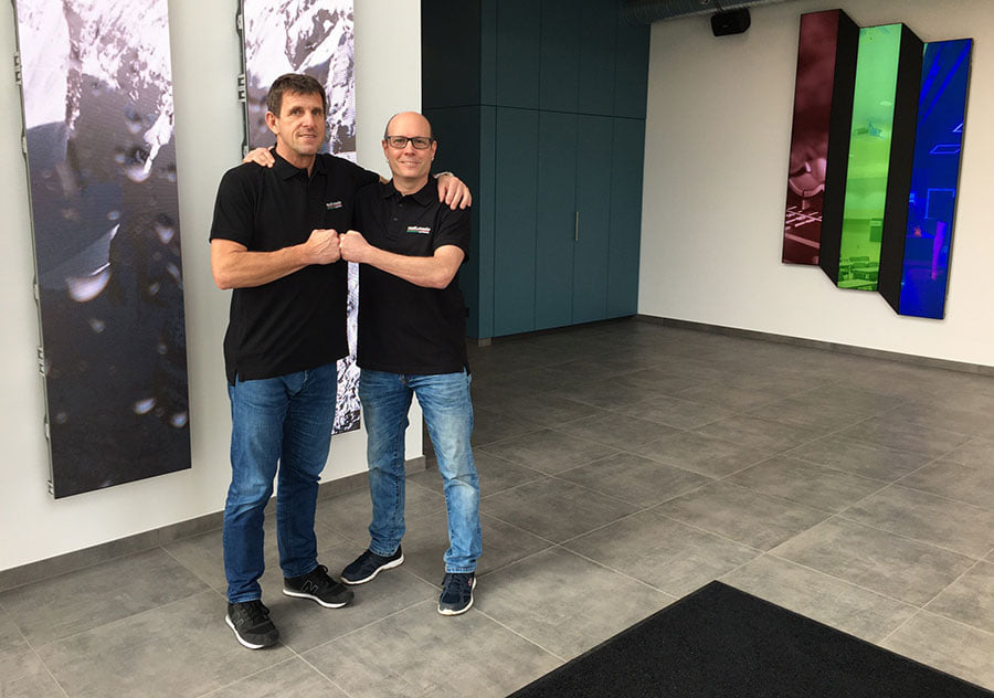 Zwei Männer in schwarzen Polohemden und blauen Jeans stehen in einem modernen Foyer, lächeln und reichen sich die Hände zu einer freundlichen Geste. Im Hintergrund sind bunte Wandkunst und große blaue Schränke zu sehen.