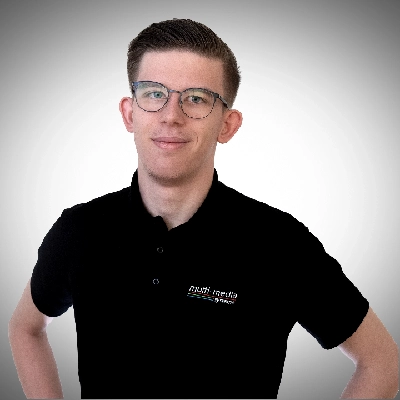 Aaron-Ries-400×400 Ein junger Mann mit kurzen braunen Haaren und Brille, der ein schwarzes Poloshirt mit einem Multimedia-Logo trägt, steht mit den Händen in den Hüften und lächelt leicht vor einem grauen Hintergrund mit Farbverlauf.
