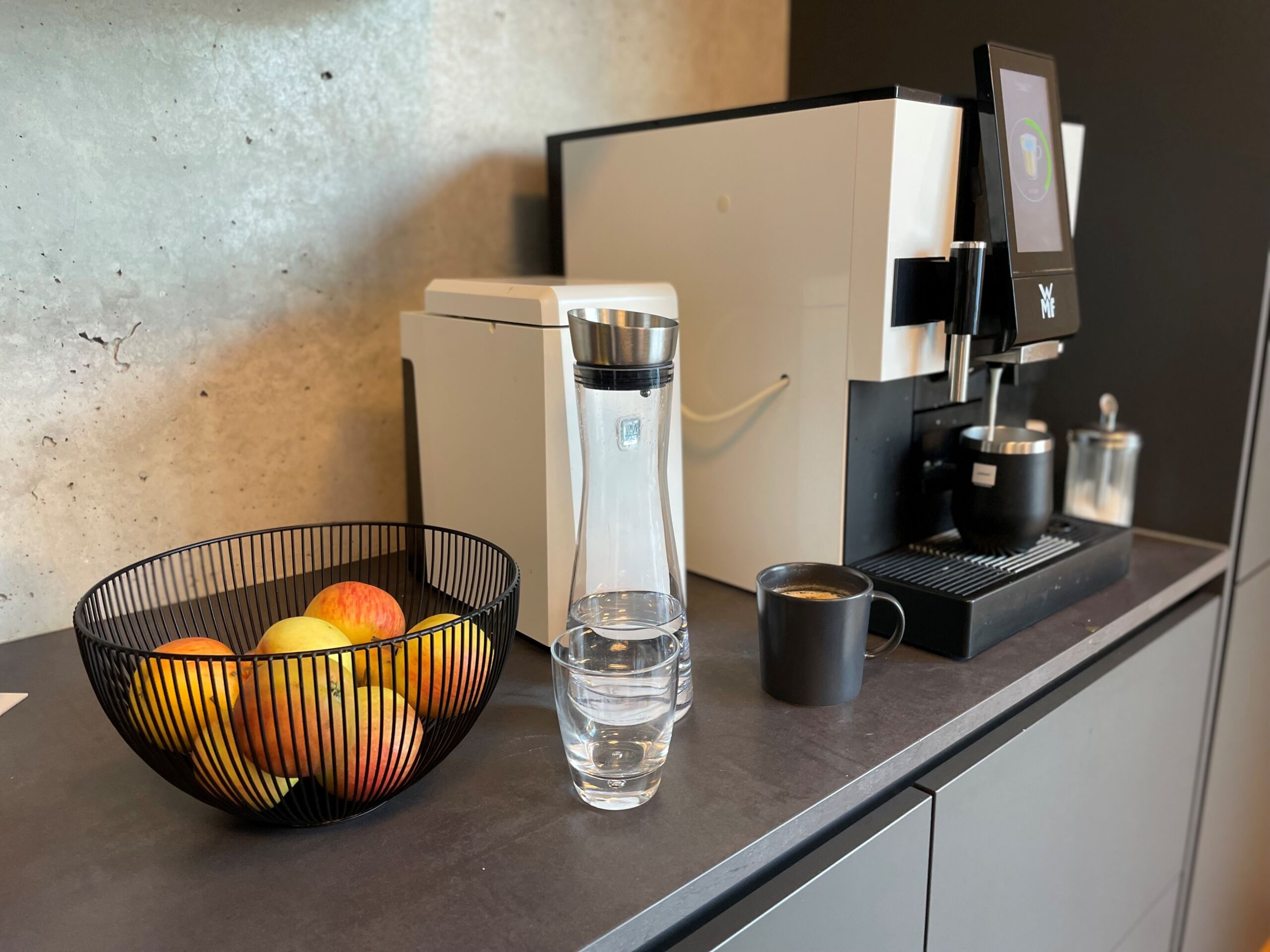 Eine moderne Küchentheke mit einer Schale Äpfel, einer hohen Glaskaraffe, einem schwarzen Kaffeebecher und einer Kaffeemaschine vor einer Betonwand.