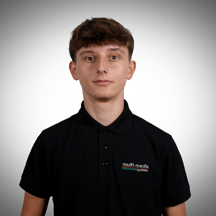 Damir-Pavelic-720×720 Ein junger Mann mit kurzen braunen Haaren, der ein schwarzes Poloshirt mit einem multi-media-Logo trägt, steht mit den Händen in den Hüften und lächelt leicht vor einem grauen Hintergrund mit Farbverlauf.