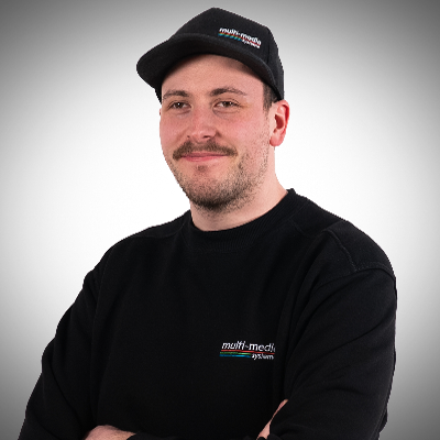 Fabian-Grieshaber-400×400 Ein Mann mit Schnurrbart, der eine schwarze Mütze und ein schwarzes Sweatshirt mit dem "Multimedia-System"-Logo trägt, steht mit verschränkten Armen und lächelt vor einem schlichten hellen Hintergrund.