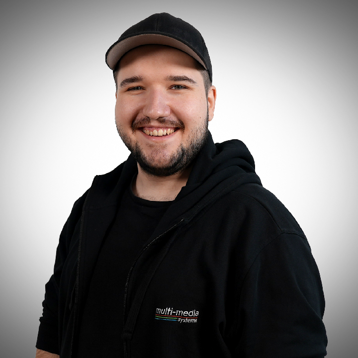 Gianni-Born-720×720 Ein junger Mann mit Bart, der eine schwarze Mütze und ein schwarzes Sweatshirt mit dem multi-media system Logo trägt, steht, lächelt vor einem schlichten hellen Hintergrund.