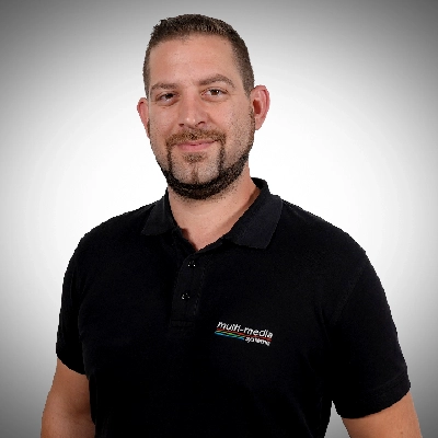 Julian-Bachmann-400×400 Mann mit dunklem Haar und Bart, der ein schwarzes Polohemd mit einem multi-media System-Logo trägt, steht vor einem schlichten, hellen Hintergrund, blickt in die Kamera und lächelt leicht.