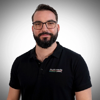 Raphael-Zippenfenig-400×400 Ein Mann mit Bart und Brille, der ein schwarzes Polohemd mit dem Logo von Multimedia Systeme trägt, steht vor einem Hintergrund mit grauem Farbverlauf, schaut in die Kamera und lächelt leicht.