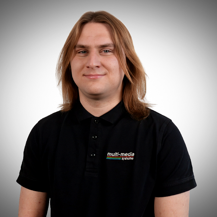 Sven-Gaidetzka-720×720 Ein junger Mann mit langem braunen Haar, der ein schwarzes Poloshirt mit einem multi-media-Logo trägt, steht mit den Händen in den Hüften und lächelt leicht vor einem grauen Hintergrund mit Farbverlauf.