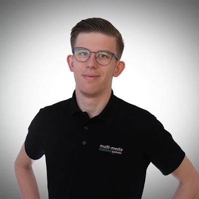 Ein junger Mann mit kurzen braunen Haaren und Brille, der ein schwarzes Poloshirt mit einem Multimedia-Logo trägt, steht mit den Händen in den Hüften und lächelt leicht vor einem grauen Hintergrund mit Farbverlauf. Ein junger Mann mit kurzen braunen Haaren und Brille, der ein schwarzes Poloshirt mit einem Multimedia-Logo trägt, steht mit den Händen in den Hüften und lächelt leicht vor einem grauen Hintergrund mit Farbverlauf.