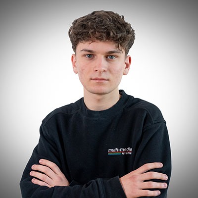 Eine junge Person mit kurzen lockigen Haaren und ernstem Gesichtsausdruck steht mit verschränkten Armen vor einem schlichten, hellen Hintergrund und trägt ein schwarzes Sweatshirt mit einem Multimedia-Logo auf der Brust. Eine junge Person mit kurzen lockigen Haaren und ernstem Gesichtsausdruck steht mit verschränkten Armen vor einem schlichten, hellen Hintergrund und trägt ein schwarzes Sweatshirt mit einem Multimedia-Logo auf der Brust.