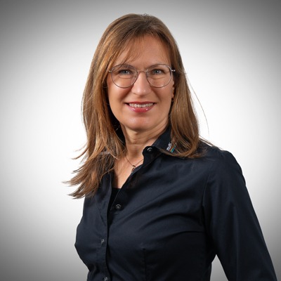 Eine Frau mit langen braunen Haaren und Brille, die ein dunkles Button-up-Hemd trägt, lächelt in die Kamera vor einem schlichten hellgrauen Hintergrund. Eine Frau mit langen braunen Haaren und Brille, die ein dunkles Button-up-Hemd trägt, lächelt in die Kamera vor einem schlichten hellgrauen Hintergrund.
