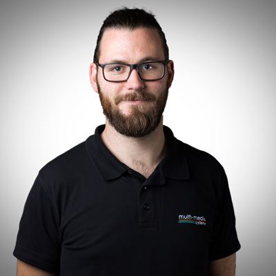 Mann mit dunklem Haar und Bart, der eine Brille und ein schwarzes Polohemd mit einem Multimedia-System-Logo trägt, steht vor einem schlichten, hellen Hintergrund, blickt in die Kamera und lächelt leicht. Mann mit dunklem Haar und Bart, der eine Brille und ein schwarzes Polohemd mit einem Multimedia-System-Logo trägt, steht vor einem schlichten, hellen Hintergrund, blickt in die Kamera und lächelt leicht.