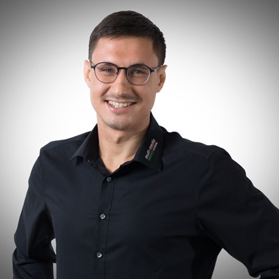 Ein lächelnder Mann mit kurzen braunen Haaren und Brille, der ein schwarzes Button-up-Hemd mit einem kleinen gestickten Logo am Kragen trägt, posiert vor einem schlichten, hellgrauen Hintergrund. Ein lächelnder Mann mit kurzen braunen Haaren und Brille, der ein schwarzes Button-up-Hemd mit einem kleinen gestickten Logo am Kragen trägt, posiert vor einem schlichten, hellgrauen Hintergrund.