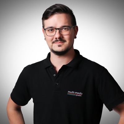 Ein Mann mit kurzen dunklen Haaren, Brille und Schnurrbart steht selbstbewusst in einem schwarzen Polohemd mit dem Logo des Multimediasystems vor einem hellgrauen Hintergrund. Ein Mann mit kurzen dunklen Haaren, Brille und Schnurrbart steht selbstbewusst in einem schwarzen Polohemd mit dem Logo des Multimediasystems vor einem hellgrauen Hintergrund.