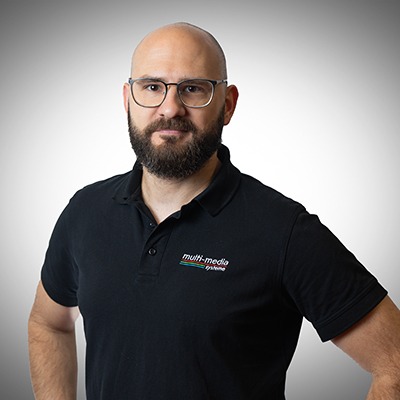 Ein bärtiger Mann mit Brille, der ein schwarzes Poloshirt von Multimedia Systeme trägt, steht vor einem schlichten, hellgrauen Hintergrund mit Farbverlauf und blickt mit neutralem Gesichtsausdruck in die Kamera. Ein bärtiger Mann mit Brille, der ein schwarzes Poloshirt von Multimedia Systeme trägt, steht vor einem schlichten, hellgrauen Hintergrund mit Farbverlauf und blickt mit neutralem Gesichtsausdruck in die Kamera.