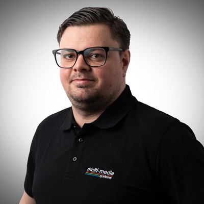 Ein Mann mit kurzen dunklen Haaren, der eine schwarze Brille und ein schwarzes Polohemd mit einem Multimedia-System-Logo trägt, steht vor einem einfarbigen Hintergrund mit hellem Farbverlauf. Ein Mann mit kurzen dunklen Haaren, der eine schwarze Brille und ein schwarzes Polohemd mit einem Multimedia-System-Logo trägt, steht vor einem einfarbigen Hintergrund mit hellem Farbverlauf.