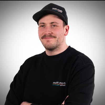 Ein Mann mit Schnurrbart, der eine schwarze Mütze und ein schwarzes Sweatshirt mit dem „Multimedia-System“-Logo trägt, steht mit verschränkten Armen und lächelt vor einem schlichten hellen Hintergrund. Ein Mann mit Schnurrbart, der eine schwarze Mütze und ein schwarzes Sweatshirt mit dem "Multimedia-System"-Logo trägt, steht mit verschränkten Armen und lächelt vor einem schlichten hellen Hintergrund.