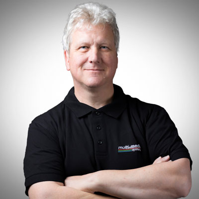 Ein Mann mit kurzen grauen Haaren, der ein schwarzes Poloshirt mit einem Multi-Med-Logo trägt, steht mit verschränkten Armen und lächelt selbstbewusst vor einem schlichten, hellen Hintergrund. Ein Mann mit kurzen grauen Haaren, der ein schwarzes Poloshirt mit einem Multi-Med-Logo trägt, steht mit verschränkten Armen und lächelt selbstbewusst vor einem schlichten, hellen Hintergrund.