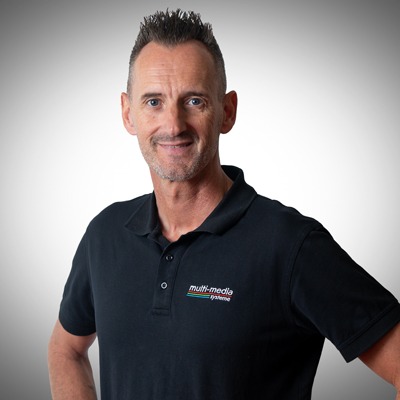 Ein Mann mit kurzen, hochgesteckten Haaren und einem schwarzen Poloshirt für Multimediasysteme steht lächelnd vor einem hellgrauen Hintergrund mit Farbverlauf. Ein Mann mit kurzen, hochgesteckten Haaren und einem schwarzen Poloshirt für Multimediasysteme steht lächelnd vor einem hellgrauen Hintergrund mit Farbverlauf.