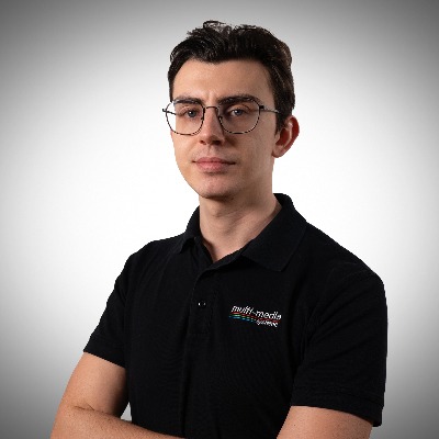 Ein junger Mann mit kurzen braunen Haaren, der eine Brille und ein schwarzes Poloshirt mit einem Multimedia-Logo trägt, steht mit verschränkten Armen vor einem schlichten, hellgrauen Hintergrund. Ein junger Mann mit kurzen braunen Haaren, der eine Brille und ein schwarzes Poloshirt mit einem Multimedia-Logo trägt, steht mit verschränkten Armen vor einem schlichten, hellgrauen Hintergrund.