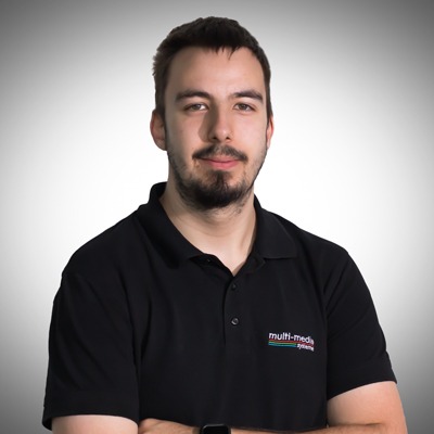 Ein Mann mit kurzen dunklen Haaren und Bart, der ein schwarzes Kragenhemd mit aufgestickten Multimediasystemen trägt, steht mit verschränkten Armen vor einem einfarbigen, hellen Hintergrund mit Farbverlauf. Ein Mann mit kurzen dunklen Haaren und Bart, der ein schwarzes Kragenhemd mit aufgestickten Multimediasystemen trägt, steht mit verschränkten Armen vor einem einfarbigen, hellen Hintergrund mit Farbverlauf.