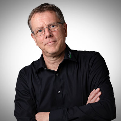 Ein Mann mittleren Alters mit kurzen Haaren und Brille, der ein schwarzes Button-up-Hemd trägt, steht mit verschränkten Armen vor einem schlichten, hellgrauen Hintergrund und schaut in die Kamera. Ein Mann mittleren Alters mit kurzen Haaren und Brille, der ein schwarzes Button-up-Hemd trägt, steht mit verschränkten Armen vor einem schlichten, hellgrauen Hintergrund und schaut in die Kamera.
