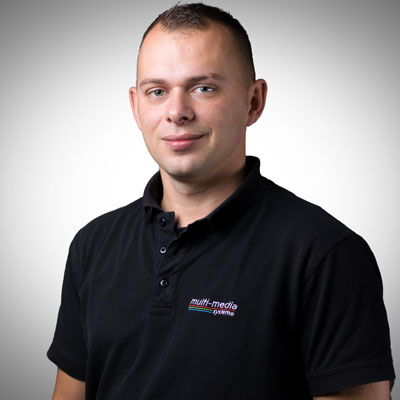 Ein Mann mit kurzen Haaren und einem schwarzen Kragenhemd mit dem Logo von Multimedia Systeme steht vor einem schlichten hellgrauen Hintergrund und lächelt sanft in die Kamera. Ein Mann mit kurzen Haaren und einem schwarzen Kragenhemd mit dem Logo von Multimedia Systeme steht vor einem schlichten hellgrauen Hintergrund und lächelt sanft in die Kamera.