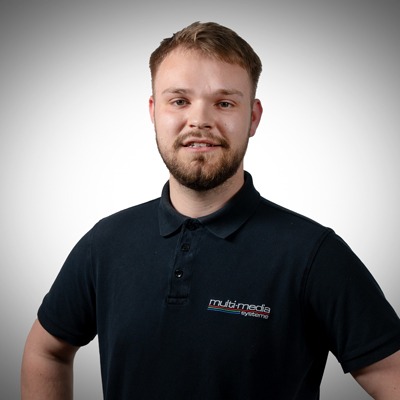 Ein junger Mann mit kurzen Haaren und Bart lächelt, trägt ein dunkles Polohemd mit einem Multimedia-Systemlogo und steht vor einem einfarbigen Hintergrund mit Farbverlauf. Ein junger Mann mit kurzen Haaren und Bart lächelt, trägt ein dunkles Polohemd mit einem Multimedia-Systemlogo und steht vor einem einfarbigen Hintergrund mit Farbverlauf.