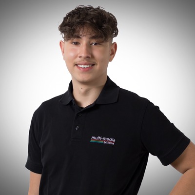 Ein junger Mann mit lockigem braunem Haar und einem leichten Schnurrbart lächelt vor einem Hintergrund mit grauem Farbverlauf. Er trägt ein schwarzes Poloshirt mit einem Multimedia-System-Logo auf der Brust. Ein junger Mann mit lockigem braunem Haar und einem leichten Schnurrbart lächelt vor einem Hintergrund mit grauem Farbverlauf. Er trägt ein schwarzes Poloshirt mit einem Multimedia-System-Logo auf der Brust.