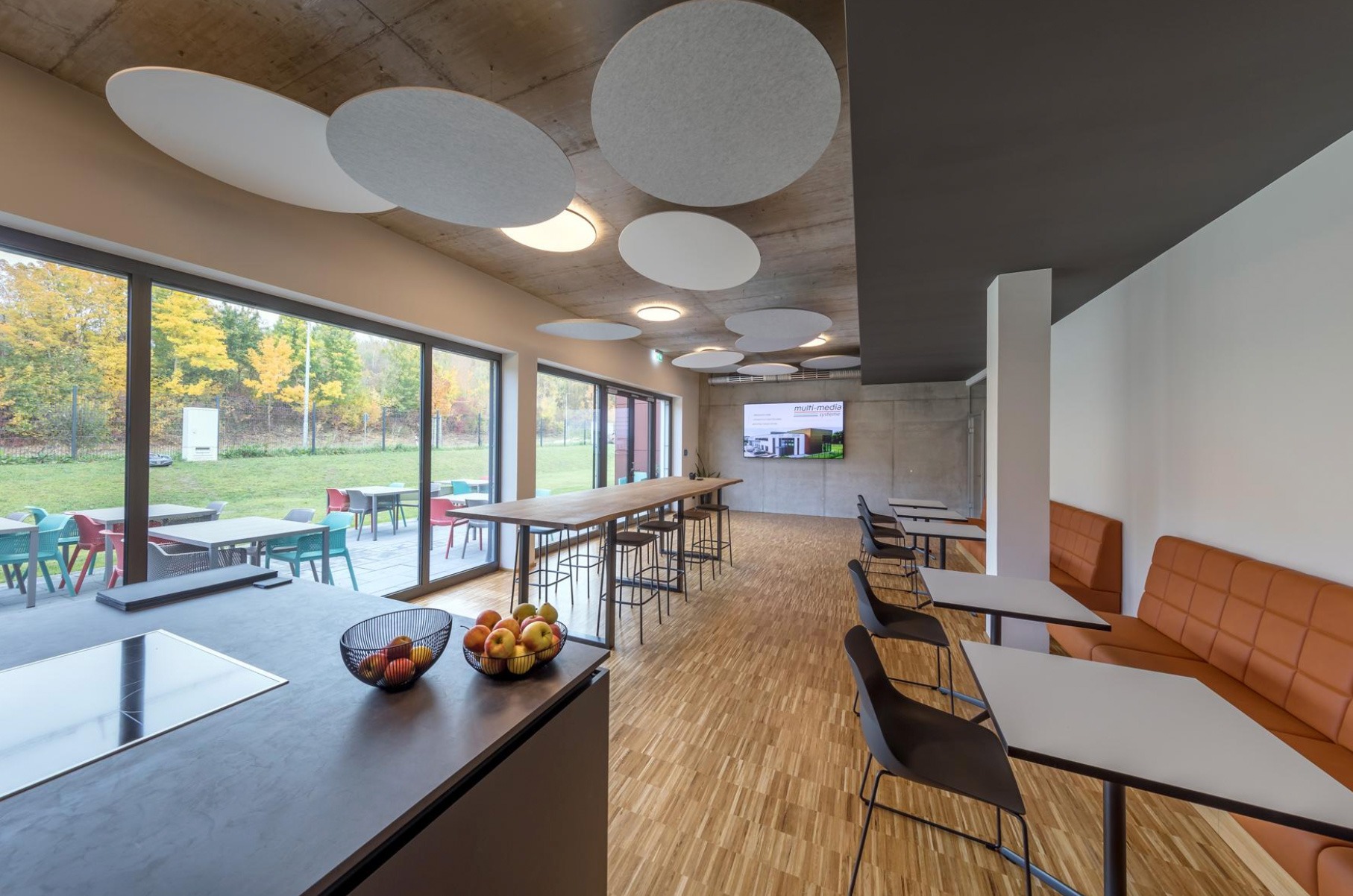 Moderne Cafeteria mit großen Fenstern, Holzboden, braunen gepolsterten Bänken, schwarzen Stühlen und Tischen. Auf der Theke steht eine Obstschale; durch die Fenster sind Sitzgelegenheiten im Freien und herbstlich gefärbte Bäume zu sehen. Der einladende Raum bietet einen inspirierenden Rahmen sowohl für Entspannung als auch für Karrieregespräche. Die Decke ist mit runden Paneelen versehen.