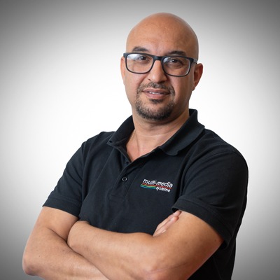 Ein Mann mit Brille, rasiertem Kopf und kurzem Bart steht mit verschränkten Armen in einem schwarzen Polohemd mit Multimedia-Logo vor einem grauen Hintergrund mit Farbverlauf. Ein Mann mit Brille, rasiertem Kopf und kurzem Bart steht mit verschränkten Armen in einem schwarzen Polohemd mit Multimedia-Logo vor einem grauen Hintergrund mit Farbverlauf.