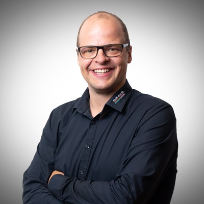 Eine lächelnde Person mit Brille und dunklem Button-up-Hemd steht mit verschränkten Armen vor einem hellen Hintergrund mit Farbverlauf. Eine lächelnde Person mit Brille und dunklem Button-up-Hemd steht mit verschränkten Armen vor einem hellen Hintergrund mit Farbverlauf.