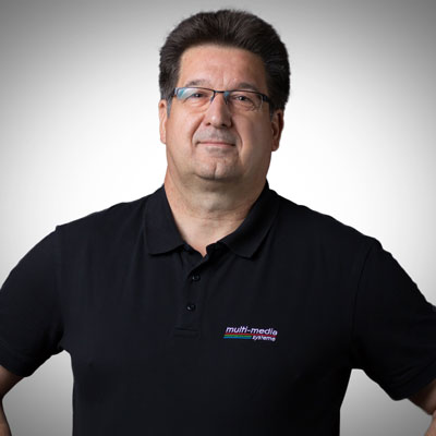 Ein Mann mit kurzen dunklen Haaren und Brille trägt ein schwarzes Poloshirt mit dem Logo von Multimedia Systems. Er steht mit einem Arm in der Hüfte vor einem schlichten, hellgrauen Hintergrund. Ein Mann mit kurzen dunklen Haaren und Brille trägt ein schwarzes Poloshirt mit dem Logo von Multimedia Systems. Er steht mit einem Arm in der Hüfte vor einem schlichten, hellgrauen Hintergrund.