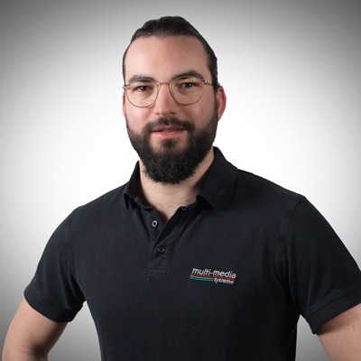 Ein Mann mit Bart und Brille, der ein schwarzes Polohemd mit dem Logo von Multimedia Systeme trägt, steht vor einem Hintergrund mit grauem Farbverlauf, schaut in die Kamera und lächelt leicht. Ein Mann mit Bart und Brille, der ein schwarzes Polohemd mit dem Logo von Multimedia Systeme trägt, steht vor einem Hintergrund mit grauem Farbverlauf, schaut in die Kamera und lächelt leicht.