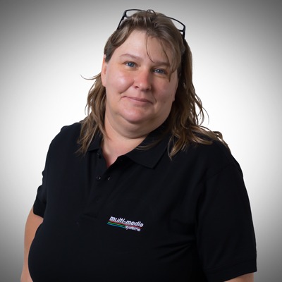 Eine Frau mit hellbraunem Haar, die eine Brille auf dem Kopf und ein schwarzes Poloshirt mit Multimediasystem trägt, steht vor einem Hintergrund mit grauem Farbverlauf und lächelt sanft in die Kamera. Eine Frau mit hellbraunem Haar, die eine Brille auf dem Kopf und ein schwarzes Poloshirt mit Multimediasystem trägt, steht vor einem Hintergrund mit grauem Farbverlauf und lächelt sanft in die Kamera.