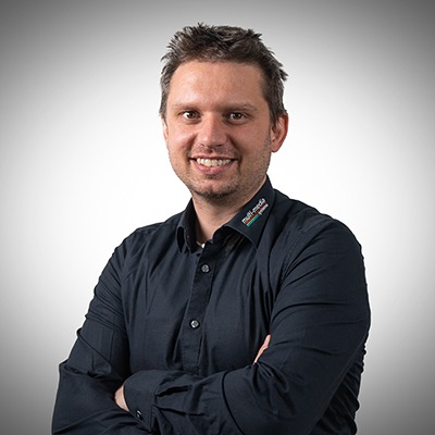 Ein Mann mit kurzen braunen Haaren und einem Bart, der ein schwarzes Button-up-Hemd mit einem Logo am Kragen trägt, steht lächelnd mit verschränkten Armen vor einem einfarbigen Hintergrund mit Farbverlauf. Ein Mann mit kurzen braunen Haaren und einem Bart, der ein schwarzes Button-up-Hemd mit einem Logo am Kragen trägt, steht lächelnd mit verschränkten Armen vor einem einfarbigen Hintergrund mit Farbverlauf.