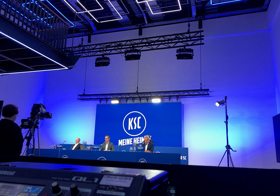 Drei Personen sitzen an einem langen Schreibtisch auf einer Bühne mit blauem Hintergrund, auf dem das KSC-Logo und MEINE HEIMAT zu sehen sind, während im Vordergrund Kameras und Beleuchtungsgeräte für Filmaufnahmen aufgebaut sind. Drei Personen sitzen an einem langen Schreibtisch auf einer Bühne mit blauem Hintergrund, auf dem das KSC-Logo und MEINE HEIMAT zu sehen sind, während im Vordergrund Kameras und Beleuchtungsgeräte für Filmaufnahmen aufgebaut sind.