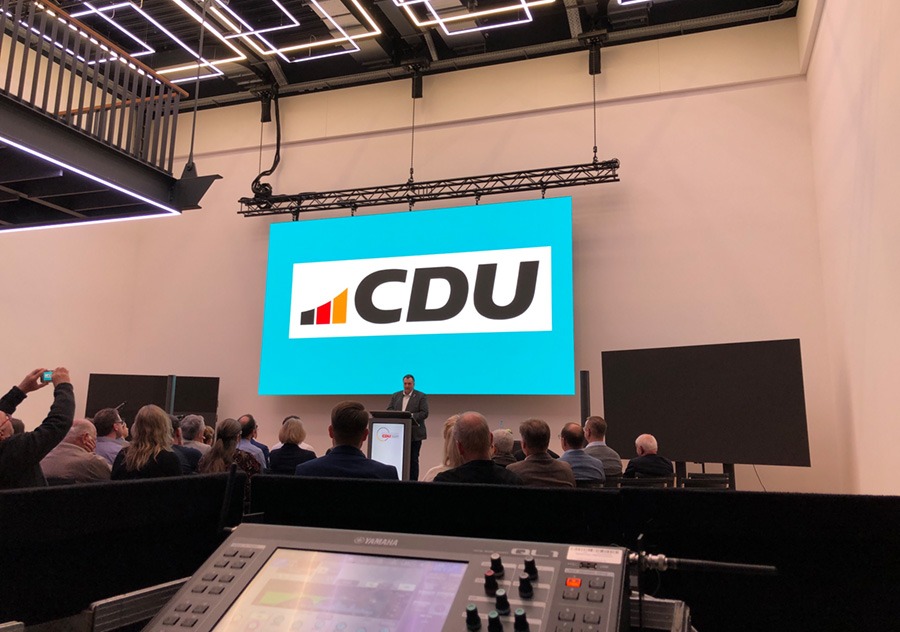 Ein Redner steht an einem Podium vor einem großen Bildschirm mit dem CDU-Logo und spricht zu einem Publikum, das in einem modernen Raum mit heller Beleuchtung und technischen Geräten im Vordergrund sitzt. Ein Redner steht an einem Podium vor einem großen Bildschirm mit dem CDU-Logo und spricht zu einem Publikum, das in einem modernen Raum mit heller Beleuchtung und technischen Geräten im Vordergrund sitzt.