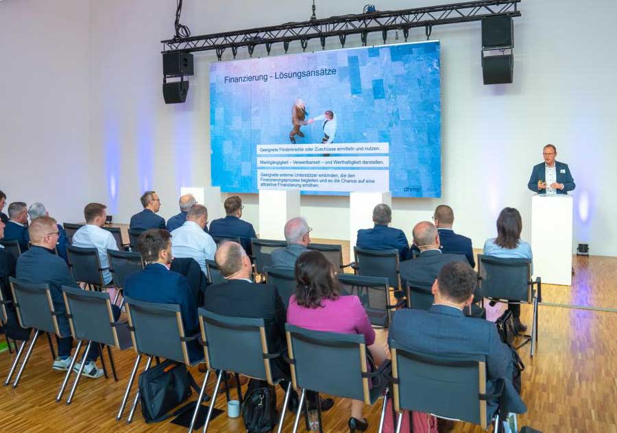 Ein Mann steht auf einem Podium und hält einen Vortrag vor einem in Reihen sitzenden Publikum; auf einem großen Bildschirm hinter ihm wird ein Dia mit dem Titel Finanzierung – Lösungsansätze und Text in deutscher Sprache angezeigt. Ein Mann steht auf einem Podium und hält einen Vortrag vor einem in Reihen sitzenden Publikum; auf einem großen Bildschirm hinter ihm wird ein Dia mit dem Titel Finanzierung - Lösungsansätze und Text in deutscher Sprache angezeigt.