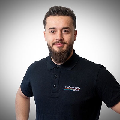 Vor einem schlichten hellen Hintergrund steht ein Mann mit kurzen lockigen Haaren und Vollbart, der ein schwarzes Poloshirt mit dem Logo von Multimedia Systems trägt und selbstbewusst in die Kamera lächelt. Vor einem schlichten hellen Hintergrund steht ein Mann mit kurzen lockigen Haaren und Vollbart, der ein schwarzes Poloshirt mit dem Logo von Multimedia Systems trägt und selbstbewusst in die Kamera lächelt.