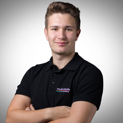 Ein junger Mann mit kurzen braunen Haaren, der ein schwarzes Polohemd mit einem Multimedia-System-Logo trägt, steht selbstbewusst mit verschränkten Armen vor einem hellgrauen Hintergrund mit Farbverlauf. Ein junger Mann mit kurzen braunen Haaren, der ein schwarzes Polohemd mit einem Multimedia-System-Logo trägt, steht selbstbewusst mit verschränkten Armen vor einem hellgrauen Hintergrund mit Farbverlauf.