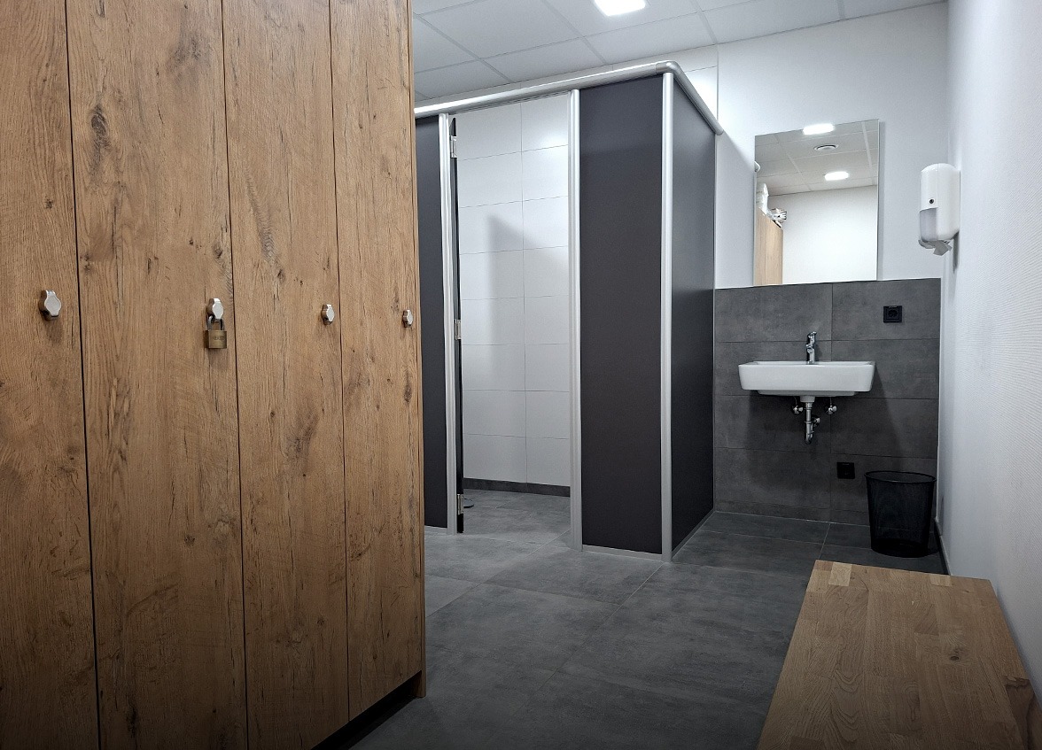 Moderner Umkleideraum mit Holzspinden, einer Bank, grauem Fliesenboden, einem Waschbecken mit Spiegel sowie einer teilweise einsehbaren Dusch- oder Toilettenkabine mit Trennwänden. Der Raum ist durchdacht gestaltet, um Sie bei Ihren Karrierezielen zu unterstützen, und verfügt über einen Seifenspender sowie einen Mülleimer, die praktischerweise in der Nähe des Waschbeckens platziert sind.