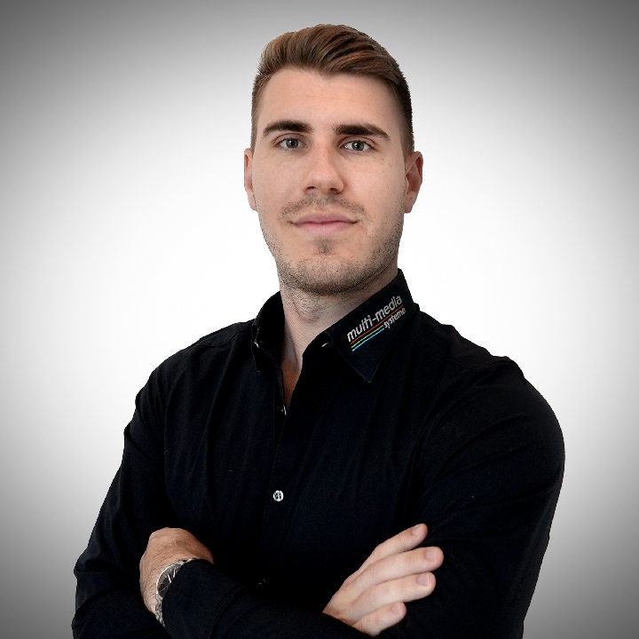 Marvin-Peterlik-720×720 Ein junger Mann mit kurzen, gestylten hellbraunen Haaren lächelt in die Kamera. Er trägt ein schwarzes Button-up-Hemd mit einem kleinen farbigen Logo am Kragen und steht vor einem schlichten, hellgrauen Hintergrund.
