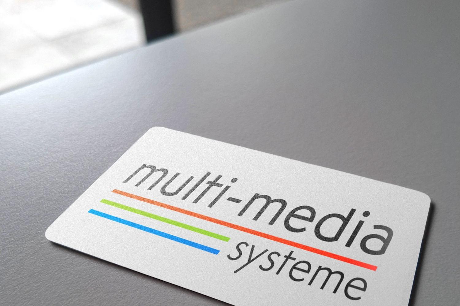 Auf einer Visitenkarte, die auf einem grauen Tisch liegt, steht „multi-media systeme“, und unter dem Wort „multi-media“ sind drei farbige Linien – blau, grün und rot – zu sehen. Das Design spiegelt einen modernen Ansatz wider, der Innovation und Karrieremöglichkeiten in der Medienbranche in den Vordergrund stellt.